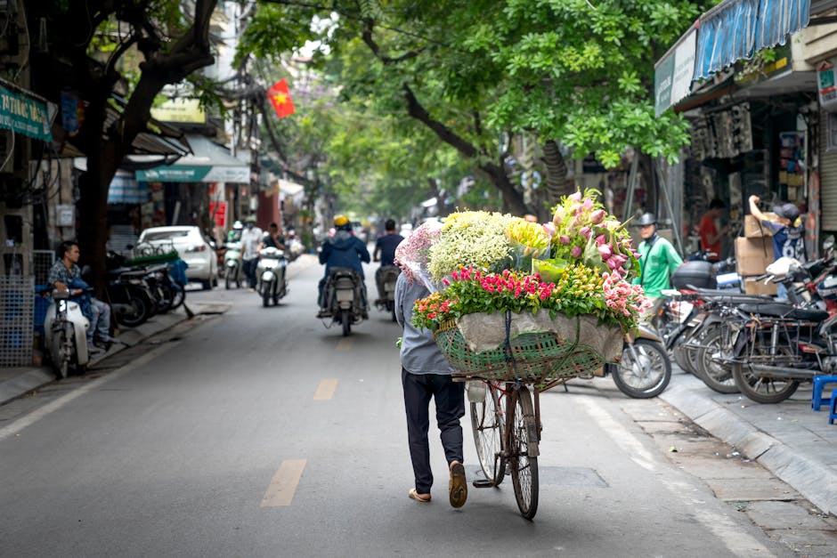 A street scene in Lee with a flower vendor pushing a bicycle cart loaded with a variety of fresh, colorful flowers including pink and white lilies, yellow and green chrysanthemums, and small pink and 