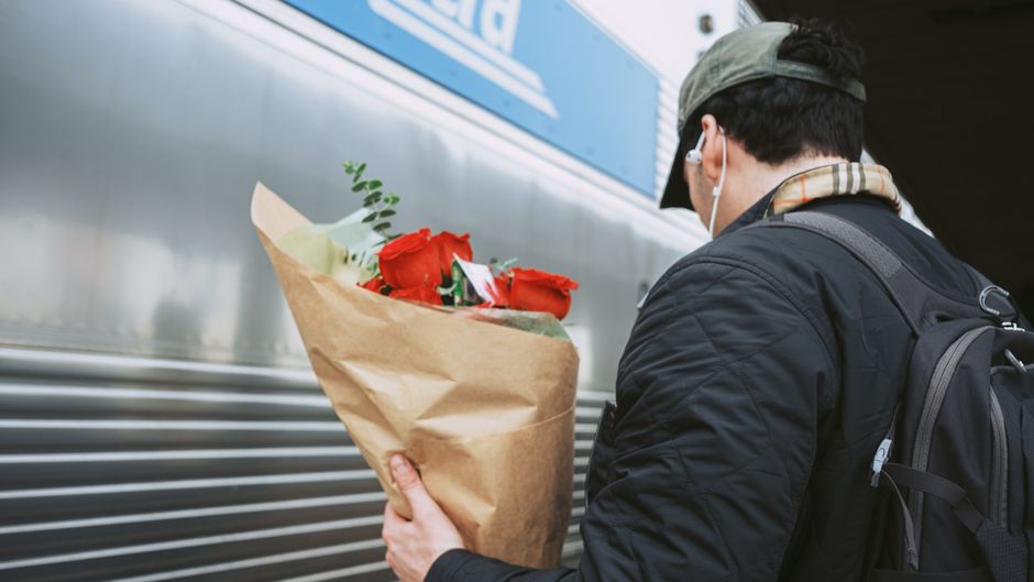 A man wearing a dark jacket, beige cap, and white earphones holds a bouquet of vibrant red roses wrapped in brown paper, standing near a silver train carriage at Lee Station. The flowers appear fresh 