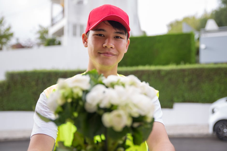 A male delivery person in a red cap and a light-colored shirt holds out a bouquet of fresh white roses and green foliage towards the camera, standing outdoors in a suburban neighborhood with neatly tr