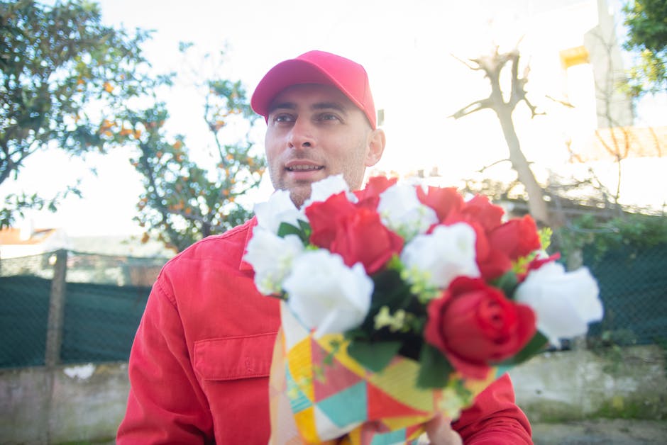 A delivery person wearing a red uniform and matching cap holds a vibrant bouquet of fresh flowers outdoors. The bouquet features red and white roses, accented with green foliage, and is wrapped in col
