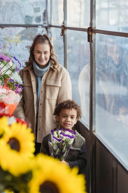 A young woman with light skin and long, wavy brown hair, smiling warmly, is standing inside a flower shop, holding a bouquet of fresh purple and green flowers. She is dressed in a beige jacket over a