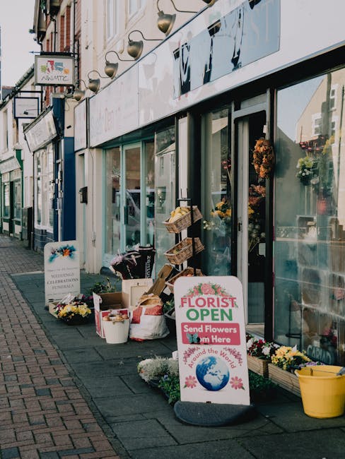 The exterior of Lee High Road flower shop features a large glass storefront displaying fresh floral arrangements inside. Outside the shop on the sidewalk, there are various flower bouquets and potted