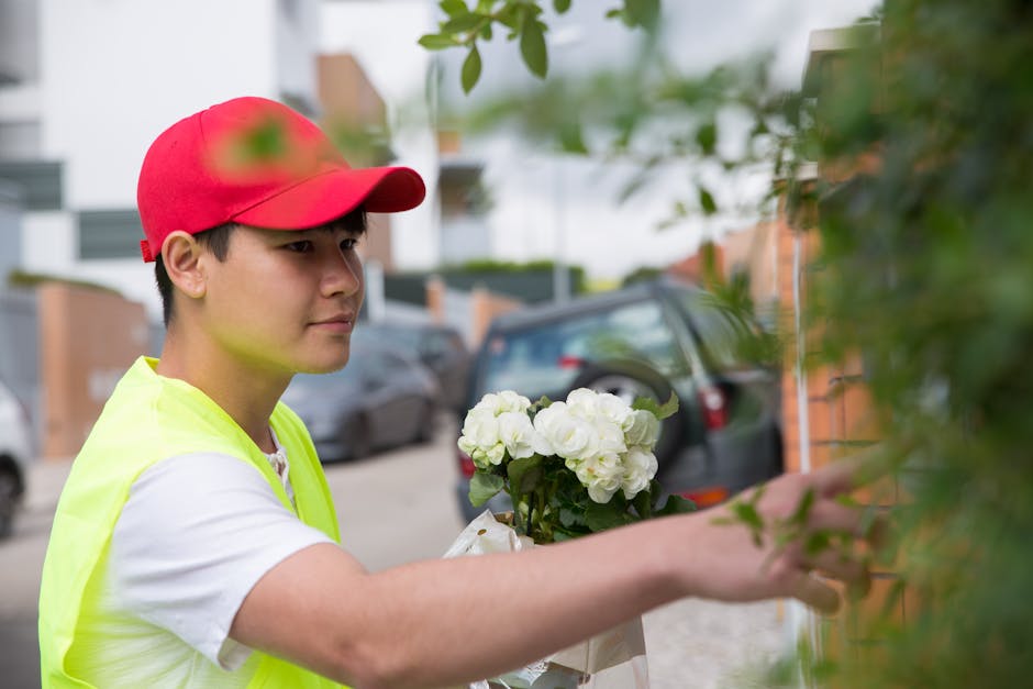 A young male florist wearing a red cap and a yellow high-visibility vest is arranging a bouquet of white roses and green foliage outside on a residential street. The arrangement is held in his left ha