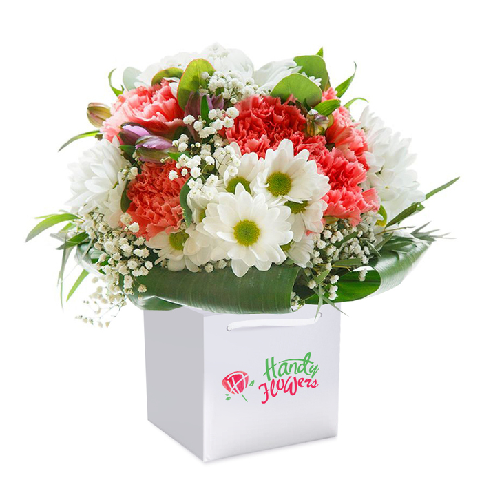 A beautifully composed bouquet showcases its bold palette and fine textures, standing upright within a crisp square white gift bag, all set against a seamless white backdrop to highlight every nuance of its artistry. At the centre, a medley of coral and peach carnations, each petal delicately rippled and soft to the eye, conveys an inviting sense of gentle luxury. Their hues transition gracefully into deeper red carnations, infusing the arrangement with energy and sophistication-a reflection of the vibrant community along Lee High Road. Sprays of freshly picked white daisies, their lime-green eyes shining like dewdrops, add a cheerful innocence, recalling sunny afternoons in Manor House Gardens. Threaded through the bouquet, clusters of pure white baby's breath bring lightness and movement, while an occasional glimpse of closed purple-tinged buds-perhaps alstroemeria ready to unfurl-promises a continual renewal, much like the seasons shifting in the parks and streets of Lee. Encircling all, lustrous, broad green leaves provide a natural frame and support, mirroring the lush gardens that define SW12's leafy character. The white bag, designed for both elegance and practicality, features sturdy ribbon handles: a detail crafted with the recipient's convenience in mind, emblematic of the service Flower Delivery Lee brings to every doorstep. Each floral element is arranged with precision, radiating freshness, abundance, and skilled familiarity with the needs of local clients. Perfect for birthdays, anniversaries, or spontaneous acts of kindness, this bouquet encapsulates the heartfelt care and expertise that set Lee's florists apart.